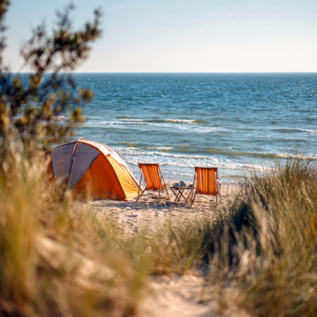 An orange camping tent and two wooden beach chairs with a small table are set up on a sandy path amidst grassy dunes, offering a serene view of the clear blue sea under a bright sky. Ideal for nature, travel, and outdoor escape themes.の素材