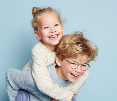 A joyful brother and sister share a piggyback ride, laughing happily together against a clean, light blue background. This image captures the bond, love, and playful spirit of siblings and family.の素材
