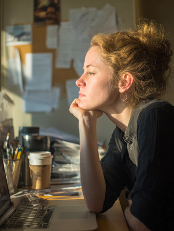 A contemplative woman rests her chin on her hand, looking away from her laptop. The warm light of the setting sun streams through the window, illuminating her face and highlighting her pensive expression as she takes a break from her work.の素材