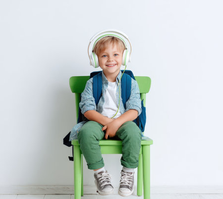 A cheerful young boy, wearing headphones and a backpack, sits on a green chair with a bright smile. He is ready for school, symbolizing the fun and excitement of learning, music, and new educational adventures.の素材