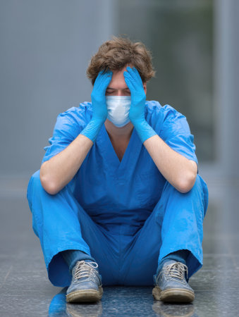 A distressed healthcare worker in a blue uniform, mask, and gloves sits on the floor with their head in their hands, conveying feelings of stress, burnout, and emotional fatigue from their job.の素材
