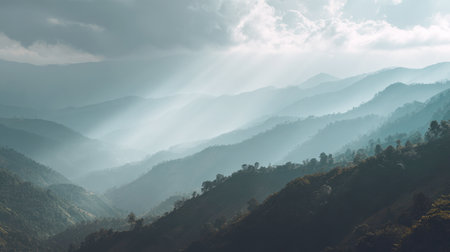 Breathtaking, horizontal landscape of a layered mountain and hill range covered in mist and fog. Intense crepuscular rays of sunlight pierce through the dramatic clouds, casting an ethereal, mystical glow over the vast, forested landscape.の素材
