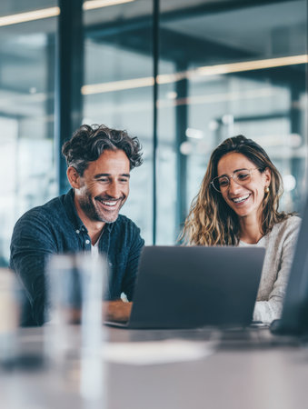 Two happy and diverse business professionals, a man and a woman, are smiling and collaborating while looking at a laptop in a bright, modern office. The image conveys teamwork, success, positive communication, and modern business solutions.の素材