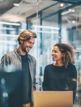 Two smiling business colleagues (one man and one woman) standing and discussing work over a laptop in a modern, glass-walled office. Represents teamwork, startup culture, collaboration, and positive professional interaction.の素材