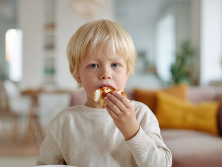 Cute portrait of a blonde toddler boy with bright eyes enjoying a piece of toast or bread with jam or butter, looking directly at the camera in a modern, bright, and comfortable home living room setting. Perfect for family, childhood, or food themes.の素材