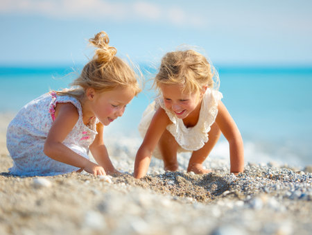 Two happy young children with blonde hair are kneeling, smiling, and digging in the sand and pebbles on a beach with clear blue ocean water. Perfect for summer, childhood, vacation, and play themes.の素材
