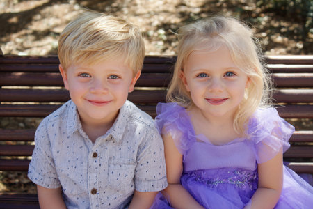Close-up, high-key portrait of two very cute young siblings (a boy and a girl) with bright blue eyes and blonde hair, sitting side-by-side on a bench and smiling at the camera, symbolizing childhood happiness, innocence, and family love.の素材