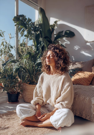 Calm Woman with Curly Hair Meditating in Cross-Legged Pose on a Jute Rug: Tranquil Indoor Setting with Large Windows, Lush Green Houseplants, and Warm Sunlight for Wellness Concept.の素材