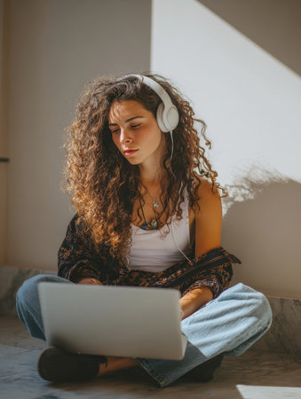 young woman with curly brown hair and freckles is sitting cross-legged against a sunlit wall, using silver laptop and listening to music or podcast through white over-ear headphones. Capturing moment of remote work, study, digital content creationの素材