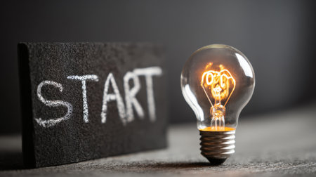 Dramatic low-key close-up of a brightly illuminated vintage filament light bulb standing next to a small chalkboard slate with the word START written in chalk, symbolizing the beginning of a new idea or project.の素材