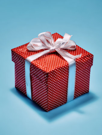 Vibrantly red gift box with white polka dots, elegantly tied with a soft silk bow and silver ribbon, positioned centrally on a bright blue background, capturing a festive, joyous, and celebratory gifting occasionの素材