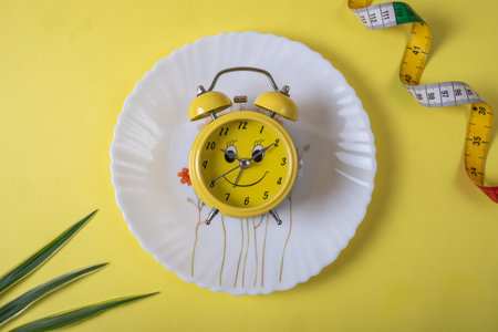 Yellow alarm clock with measuring tape on yellow background. Diet concept.の写真素材