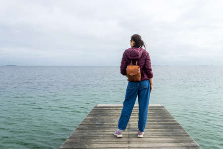 Girl with a backpack, looks at the sea, on a wooden pier. Back view. Freedom. Trips. Lifestyle.の写真素材