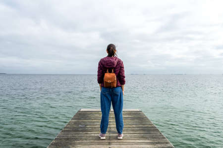 Young girl with a backpack, looks at the sea, on a wooden pier. Freedom. Trips. Lifestyle.の写真素材