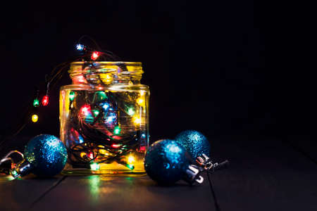 A glowing garland in a glass jar and Christmas decorations on a dark wooden background. New Year, Christmas postcard. Copy space. Holiday backgroundの写真素材