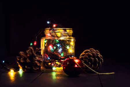 A glowing garland in a glass jar and Christmas decorations on a dark wooden background. New Year, Christmas postcard. Copy space. Holiday backgroundの写真素材