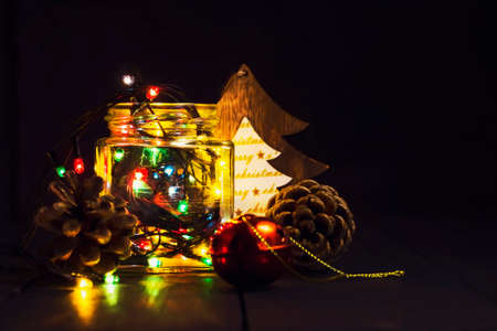 A glowing garland in a glass jar and Christmas decorations on a dark wooden background. New Year, Christmas postcard. Copy space. Holiday backgroundの写真素材