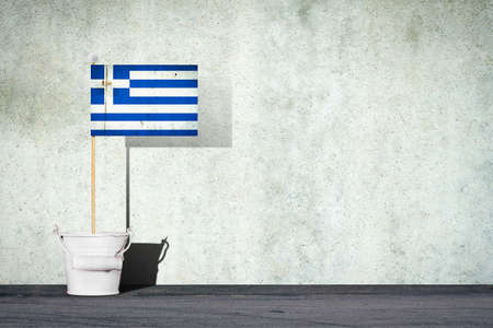 Flag of Greece, on a stick, in a small bucket, against the background of a concrete wall. Copy space. Signs and Symbols. Flags.の写真素材