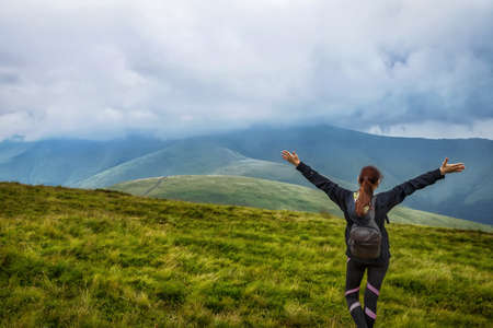 Girl with a backpack and raised hands in the mountains. Low clouds. Fog. Achievement of the goal. Success. Tourism. Travels.の写真素材