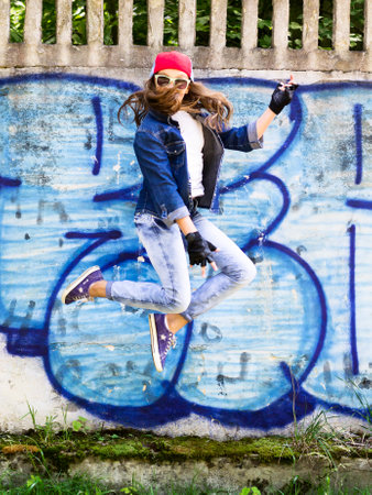 Cute young blonde teenager girl in a baseball cap and jeans shirt jumping against a stone wall background. Hip hop,の写真素材