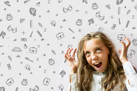 Concept Back to school. A sweet, green-eyed, fair-haired, smiling girl with hands up, against the wall with various school supplies, hand-drawn. Background. Education.の写真素材