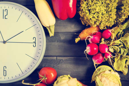 Clock and fresh vegetables. Zucchini, bell peppers, carrots, cabbage, cauliflower, radish, lettuce, tomato. Concept of a healthy diet Copy space Time to changeの写真素材