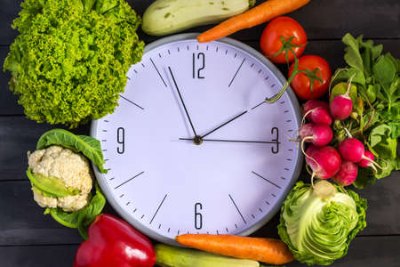 Clock and fresh vegetables. Zucchini, bell peppers, carrots, cabbage, cauliflower, radish, lettuce, tomato. Concept of a healthy diet Copy space Time to changeの写真素材