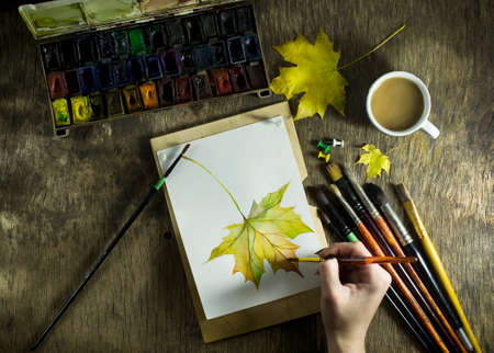 The hand of a young girl which draws a sketch of an autumn leaf. With a cup of coffee on the wooden table. Top viewの写真素材