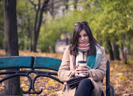 Young beautiful girl with a coffee in a paper cup. Against the background of the autumn parkの写真素材