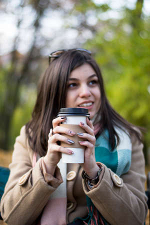 Young beautiful girl with a coffee in a paper cup. Against the background of the autumn parkの写真素材