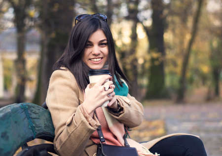Young beautiful girl with a coffee in a paper cup. Against the background of the autumn parkの写真素材