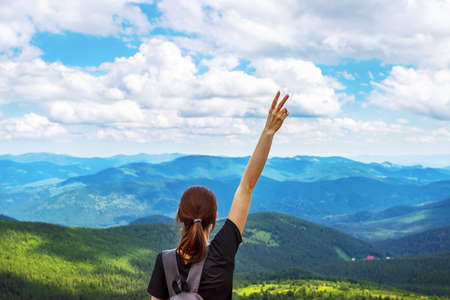 A young girl with a backpack, with raised hands, and two fingers up on a background of beautiful mountains, and a beautiful cloudy sky. Leisure. Hiking. Healthy lifestyle.の写真素材