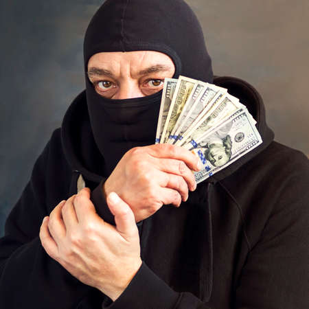Thief in balaclava with dollars in hand. Corruption. Bribe. Fraud. Dark backgroundの写真素材