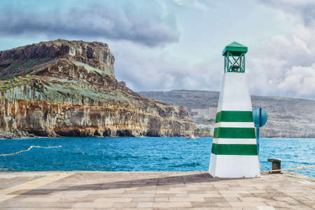 Small lighthouse on the quay. against the background of the ocean, beautiful mountains and a cloudy sky. Travels. Objectsの写真素材