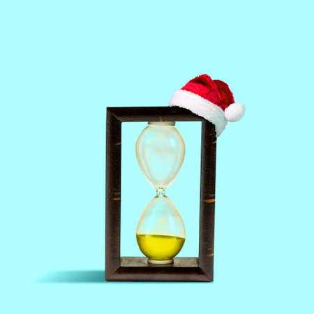 Hourglass in Santa Claus Hat. Isolated on light blue background. The symbol of the outgoing year. Festive background.の写真素材