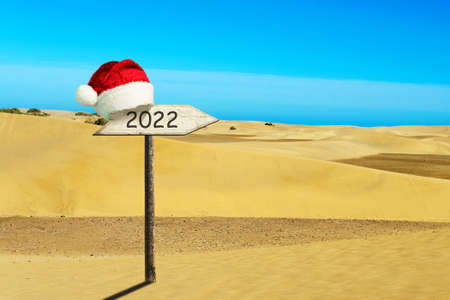 2022, a wooden sign in a Santa Claus Hat on a background of sand dunes. New Year card. Festive...
