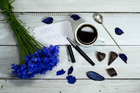 Blue wildflowers with a cup of coffee and candies on a white wooden tableの写真素材