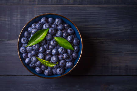 Blueberries with green leaves in a bowl, on a dark blue wooden background. Healthy food. Diet. Top view. Berries.の写真素材