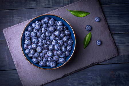 Blueberries with green leaves in a bowl, on a stone stand, on a dark blue wooden table. Healthy food. Diet. Top view. Berries.の写真素材