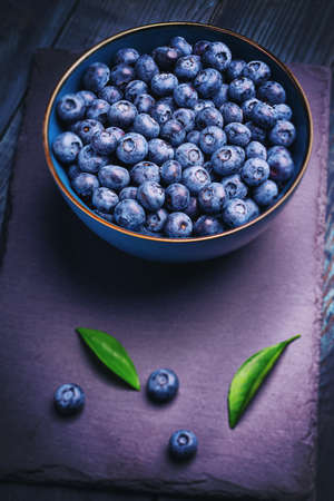 Blueberries with green leaves in a bowl, on a stone stand, on a dark blue wooden table. Healthy food. Diet. Top view. Berries.の写真素材