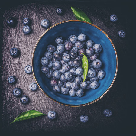 Blueberries with green leaves in a bowl, on a stone stand, on a dark blue wooden table. Toned. Healthy food. Diet. Top view. Berries.の写真素材
