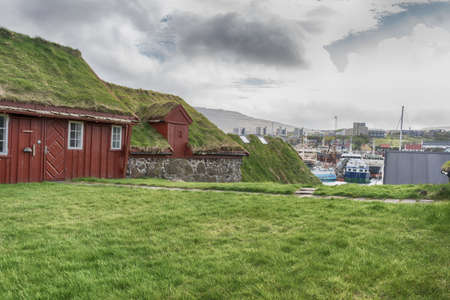 Beautiful wooden, red, small houses with a grass roof, against the backdrop of a harbor with ships. Faroe islands. Denmark. Architecture.の写真素材