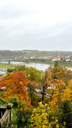 Bright autumn leaves in various shades of orange and yellow frame the view of a calm river. Soft gray skies hang above the valley, adding to the peaceful atmosphere of the landscape.の写真素材