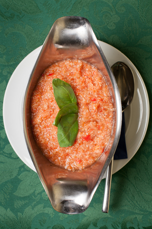 Gazpacho soup garnished with basil leaves in a silver terrine with spoon on white plate and green brocade tablecloth backgroundの写真素材