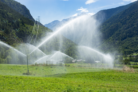 sprinkler system watering mountain pasture, tree-covered mountains in background, sunny day, mist hanging over fieldの写真素材