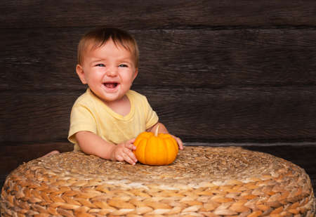 cute baby with a small orange pumpkin smiling near a straw table on a wooden backgroundの写真素材