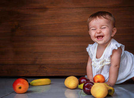 happy cute baby girl crawling on the floor with fruits apples, plums, bananas, mango on a wooden backgroundの写真素材