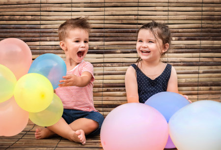 two children with balloons on the wooden background. boy and girl sit on bamboo background laughing...