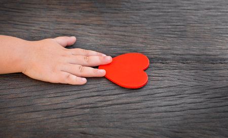 child hand reaches for red heart on dark wooden backgroundの写真素材
