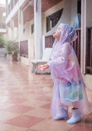 A girl in a raincoat in the rain. Child catches raindrops with hands and tongueの写真素材